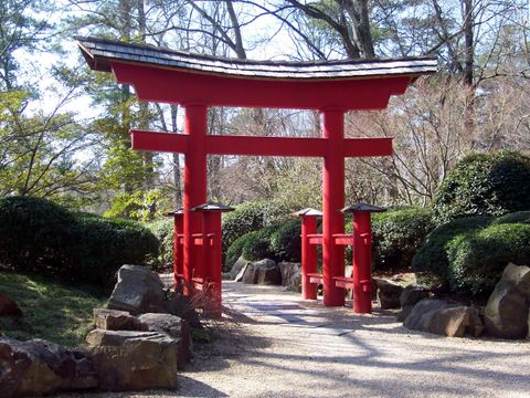 A vermilion torii with a shingled gabled roof and supporting side posts, on a gravel path through a Japanese garden with boulders and clipped shrubs.