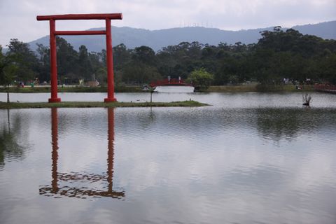 A vermilion torii standing on a grassy islet in a lake, reflected in the still water, with a red arched footbridge, walking paths, and forested hills behind it.