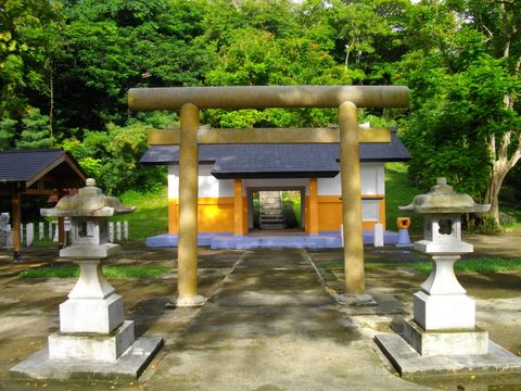 A tan stone torii with thick cylindrical pillars on a paved path, flanked by a pair of stone lanterns, with a small white and orange shrine building behind it against a forested hillside.