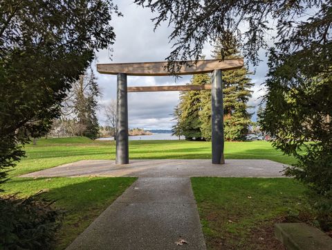 A minimalist modern torii of two grey basalt pillars supporting two horizontal cedar beams, on a paved plaza overlooking a lake with evergreens behind.