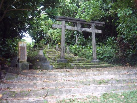 A weathered grey stone torii atop moss-covered stone steps, framed by dense tropical foliage, with an inscribed stone stele and a small stone monument to the left.
