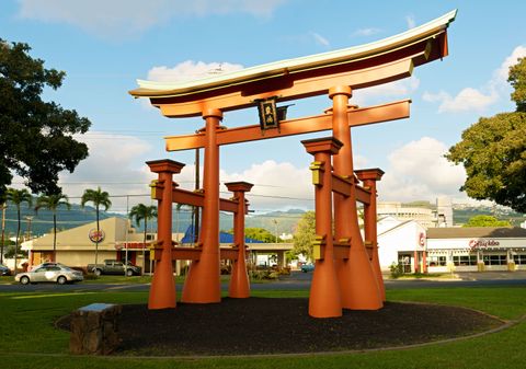 A large orange torii with a sweeping upturned roof, each pillar braced by clusters of splayed support posts, standing on a grassy traffic island with palm trees, green hills, and a strip of commercial buildings behind it.