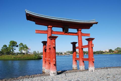 A vermilion torii with a curved blue-grey roof standing on the rocky shore at the edge of Epcot's World Showcase Lagoon.